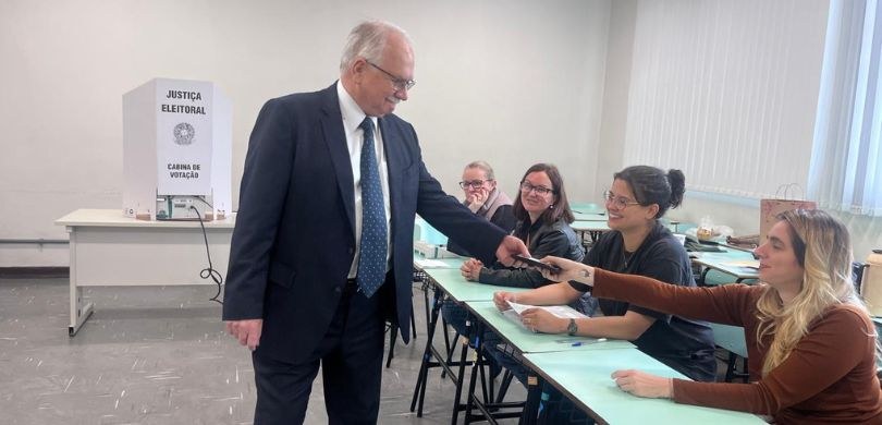 Foto em que um homem de terno escuro está em pé, entregando o celular a uma das quatro component...