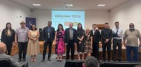 Fotografia de 12 pessoas, cinco mulheres e sete homens, posando sorrindo em um auditório. Ao fun...