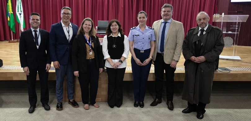 Foto em que aparecem três mulheres e quatro homens posando em frente ao palco do auditório do TR...