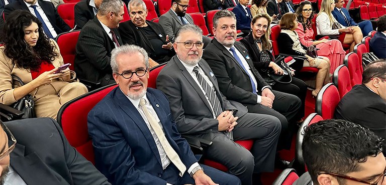 Fotografia de várias pessoas sentadas em cadeiras vermelhas em um auditório. O foco da imagem sã...