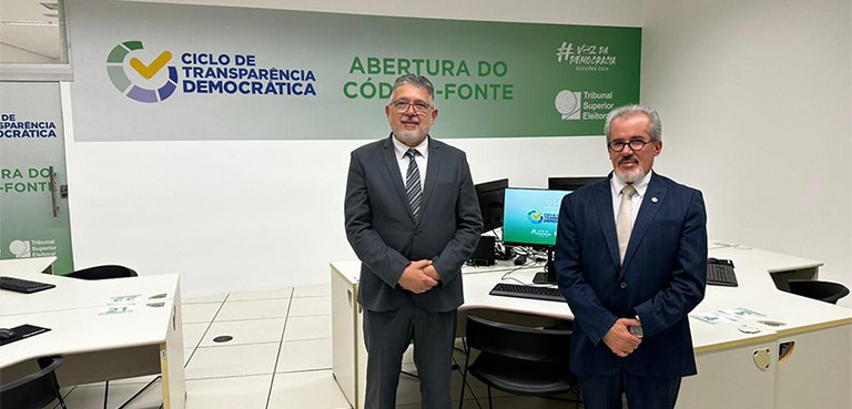 Fotografia de dois homens de terno posando sorrindo em uma sala branca. Ao fundo, há mesas branc...