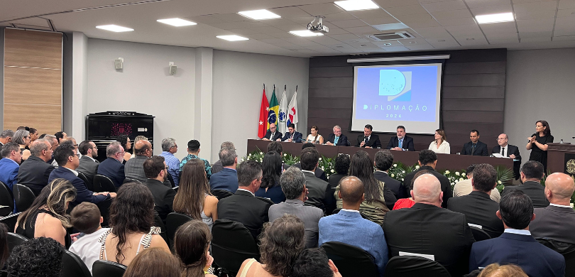 Fotografia de uma mesa de autoridades, composta por homens e mulheres, em um palco de auditório....