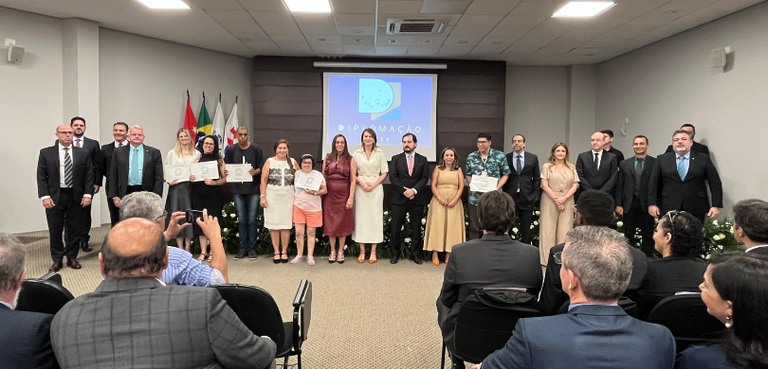 Fotografia de várias pessoas, entre homens e mulheres, em pé em um auditório. Algumas delas segu...