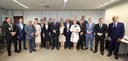 Foto de um grupo de 16 homens e cinco mulheres, em pé, em uma sala de reuniões, olhando para a c...