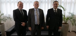 Fotografia de três homens de terno posando sorrindo. Ao fundo, há um sofá preto, dois vasos de p...