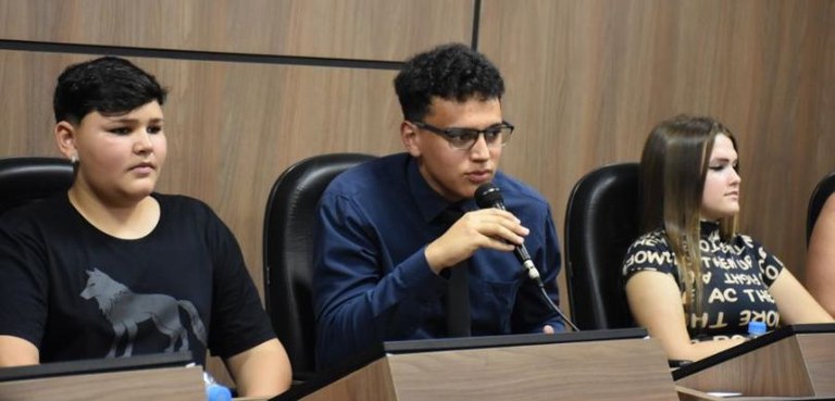 Fotografia de dois meninos e uma menina sentados em cadeiras com estofado preto atrás de púlpito...