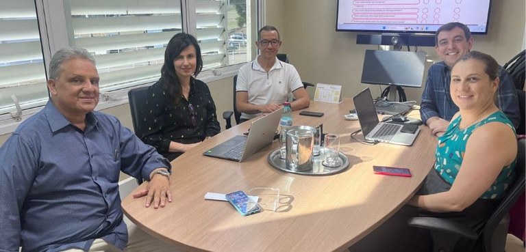 Foto em que aparecem duas mulheres e três homens posando para a foto em volta de uma mesa elípti...