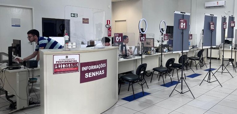 Fotografia de uma sala com paredes brancas, com quatro guichês de atendimento numerados. Cada um...