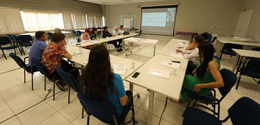 Fotografia de diversas pessoas sentadas em cadeiras com estofado azul de frente para carteiras b...