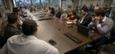 Fotografia de diversas pessoas, entre homens e mulheres, ao redor de uma mesa de reunião em made...