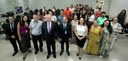 Fotografia de diversas pessoas, entre homens e mulheres, posando sorrindo em uma sala com cadeir...