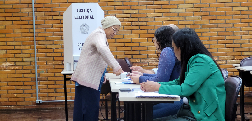 Fotografia de duas mesárias recebendo uma eleitora idosa na mesa de votação. Ao fundo, observa-s...