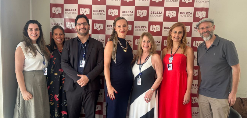 Fotografia de dois homens e cinco mulheres posando sorrindo em frente a um banner branco e verme...