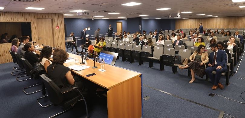 Fotografia de um auditório com homens e mulheres sentados em cadeiras com estofado cinza-claro. ...