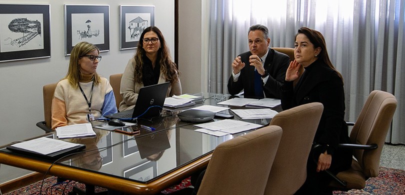 Fotografia de um homem e três mulheres sentados em cadeiras de estofado bege ao redor de uma mes...