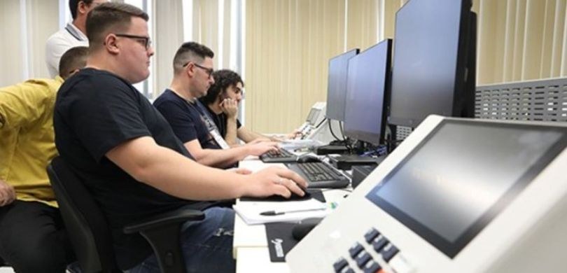 Fotografia de quatro homens sentados em cadeiras pretas diante de computadores pretos, enquanto ...