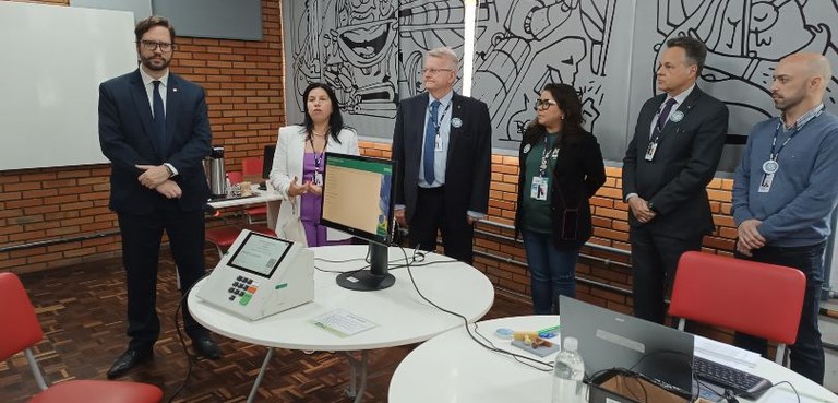 Foto em que aparecem duas mulheres e quatro homens sem pé diante de duas mesas brancas, sobre as...