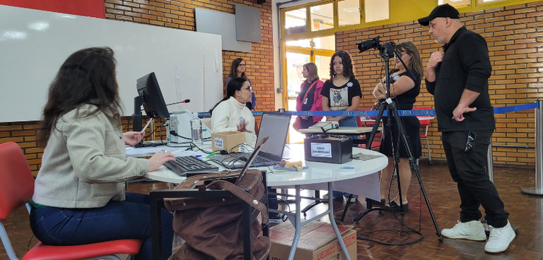 Fotografia de duas mulheres sentadas ao redor de uma mesa redonda branca, sobre a qual há comput...