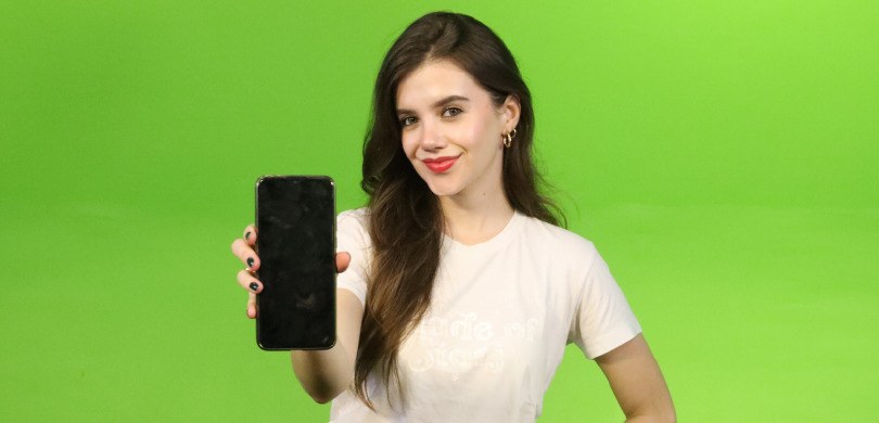 Fotografia de uma jovem mulher sorrindo e segurando um smartphone com a mão direita em direção à...