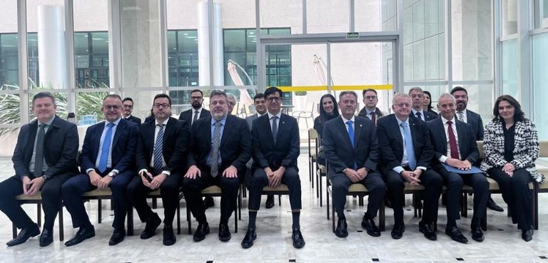 Fotografia de diversas pessoas, a maioria homens de terno, posando sorrindo. Eles estão sentados...