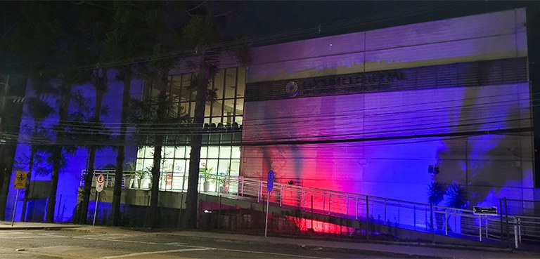 A fotografia é da fachada do Fórum Eleitoral à noite. O prédio está iluminado nas cores vermelho...