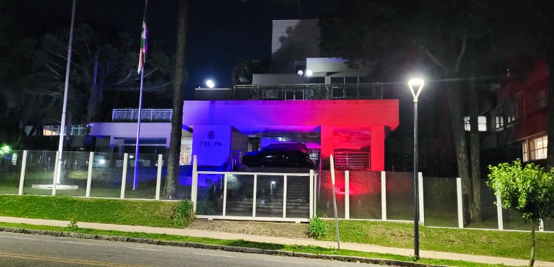 A fotografia é da fachada do edifício-sede do TRE-PR à noite. Há um muro de vidro por toda exten...