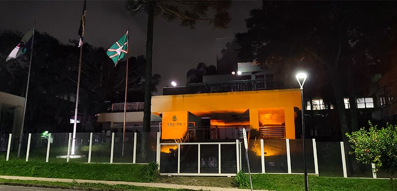 Foto da fachada do edifício-sede do TRE-PR iluminado com luz cor de laranja. Do lado esquerdo da...