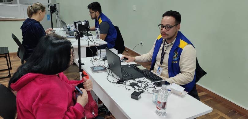 Dois servidores da Justiça Eleitoral, com colete azul e amarelo, atendem duas eleitoras.