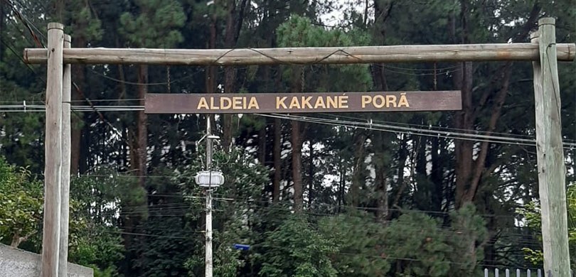 Fotografia da entrada da aldeia Kakané Porã. Ao fundo, há árvores e um poste.

