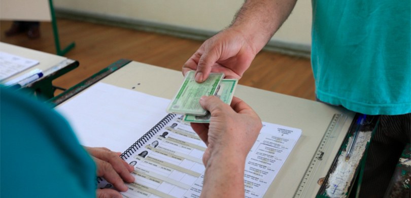 Fotografia de uma pessoa entregando documentos para votar. Abaixo das mãos do eleitor tem o cade...