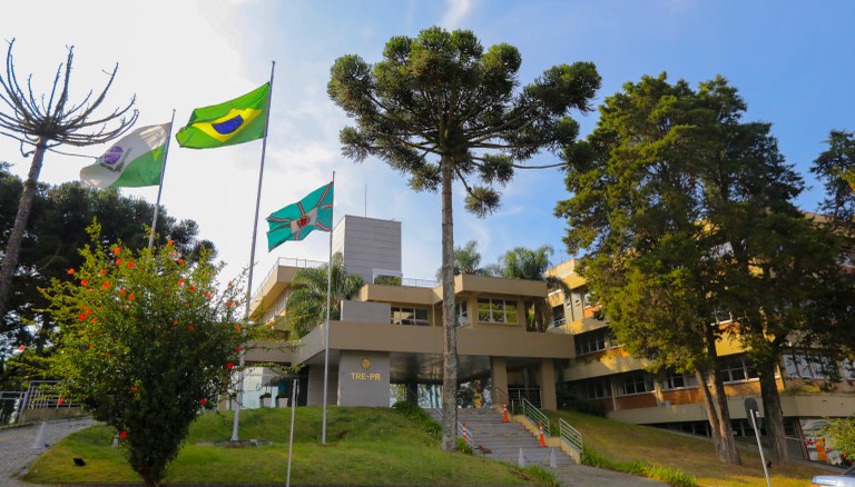 Foto da fachada do edifício-sede do TRE-PR em dia ensolarado. Na foto veem-se árvores em frente ...