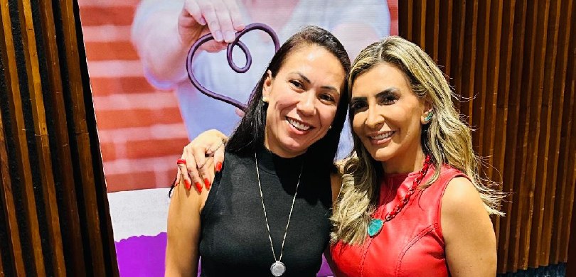 Fotografia de duas mulheres posando sorrindo abraçadas. Uma delas tem cabelos escuros e veste um...