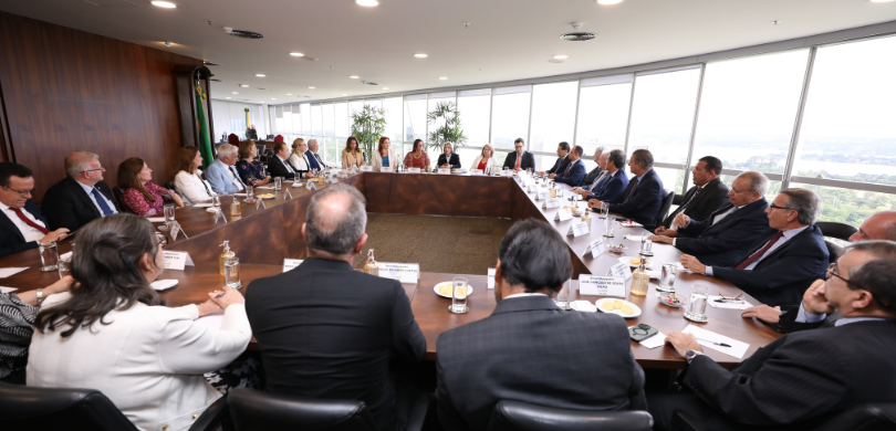 Fotografia de cerca de 30 pessoas, entre homens e mulheres, sentadas ao redor de uma mesa de reu...