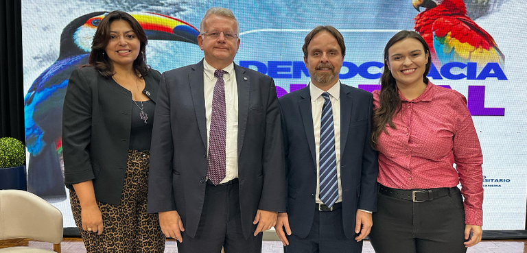 Fotografia de dois homens de terno e duas mulheres, que vestem roupas sociais, posando sorrindo....