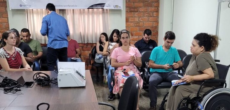 Fotografia de uma sala em que várias pessoas, entre homens e mulheres, aparecem sentados em cade...