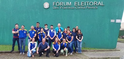 Fotografia de homens e mulheres vestindo coletes da Justiça Eleitoral na cor azul, com detalhes ...