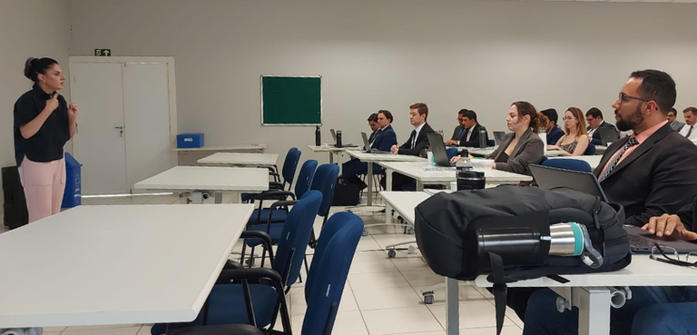 Fotografia de uma mulher, que veste camisa preta e calça rosa-clara, falando diante de uma plate...