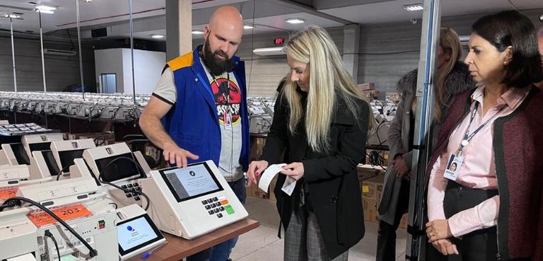 Foto em que aparecem, em primeiro plano, três pessoas em torno de uma urna eletrônica sobre uma ...