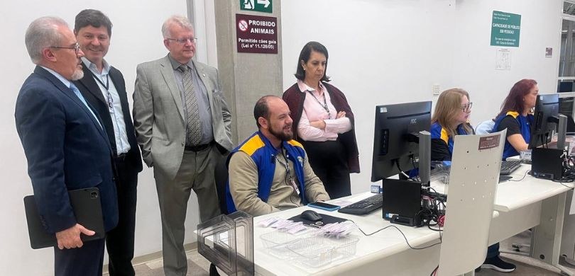 Foto em que aparece em primeiro plano um homem vestindo colete azul e amarelo da Justiça Eleitor...