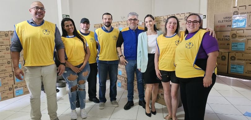 Em uma sala com paredes brancas, quatro homens e quatro mulheres, que trajam coletes amarelos co...