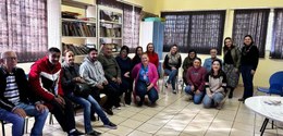 Fotografia de homens e mulheres sentados em cadeiras próximas às paredes de uma sala. A sala tem...