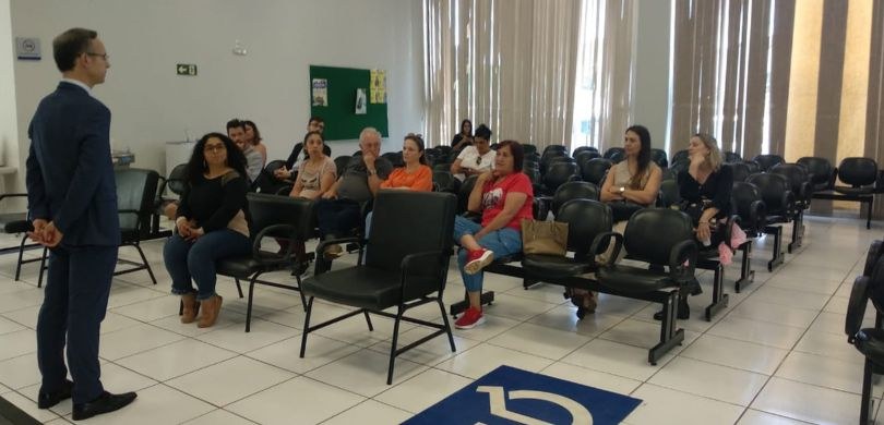 Fotografia de um homem que veste terno azul-escuro falando para pessoas sentadas em cadeiras ali...