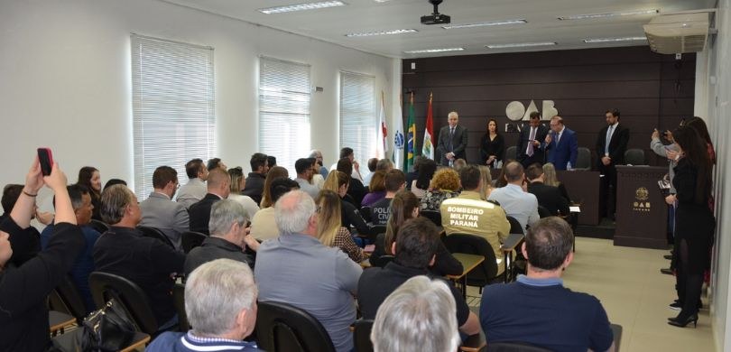 Foto de um auditório em que se veem, em primeiro plano, pessoas sentadas de costas em um pequeno...