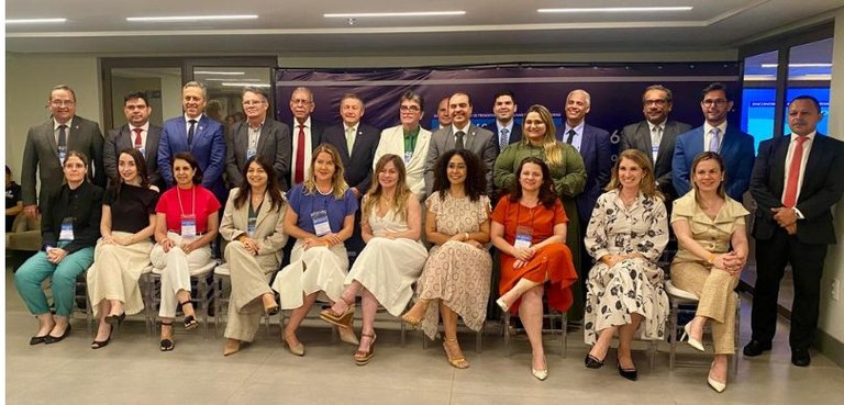 Em frente a duas portas de vidro e um painel, várias pessoas posam para a foto. As mulheres estã...